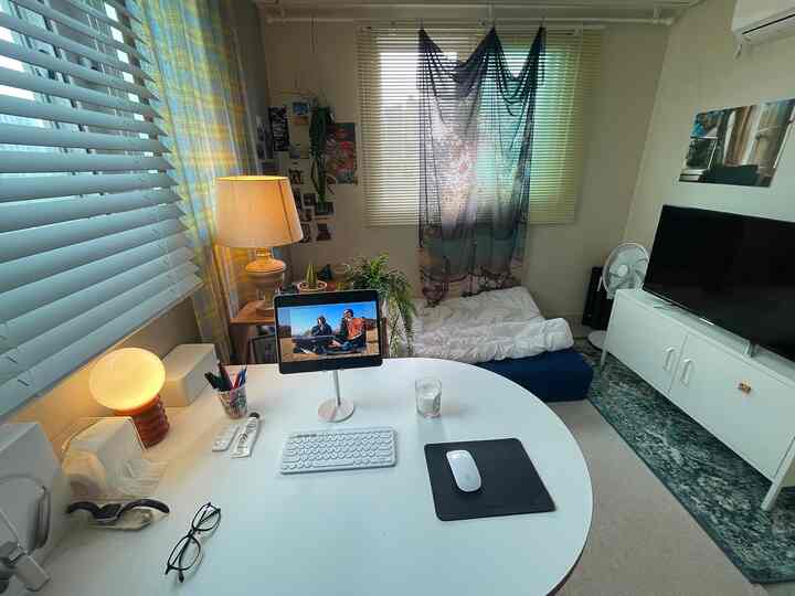 白とナチュラルな色調の一人暮らしの部屋、ホームオフィスの机、ベッド、カーテン、テレビ台が配置された雰囲気のあるインテリア