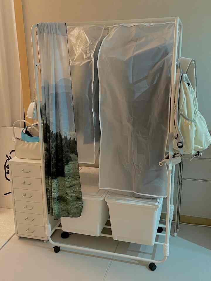 白を基調とした狭い部屋に移動式ハンガー、ドレッサー、リビングボックスが配置された実用的な収納空間