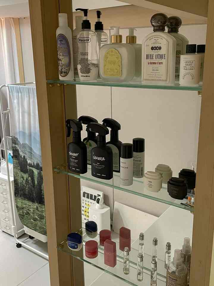 ナチュラルカラーのバスルームの化粧台、ガラス棚に多様なボディローションとスプレーが整然と並ぶ清潔な空間