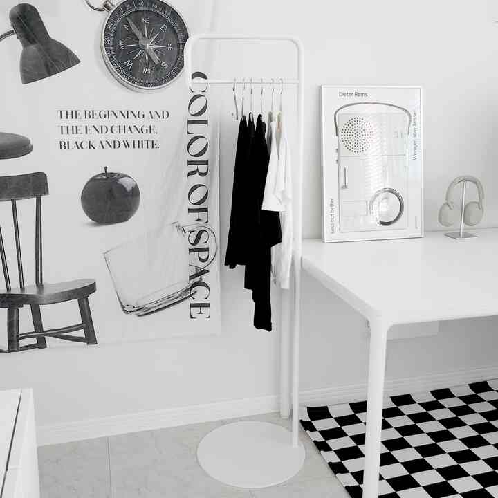 モノトーンのホワイトを基調としたワークスペース、左側の壁ポスターとコートラック、右側のデスクとチェッカーフラッグ柄ラグが配置されたシンプルな雰囲気のインテリア