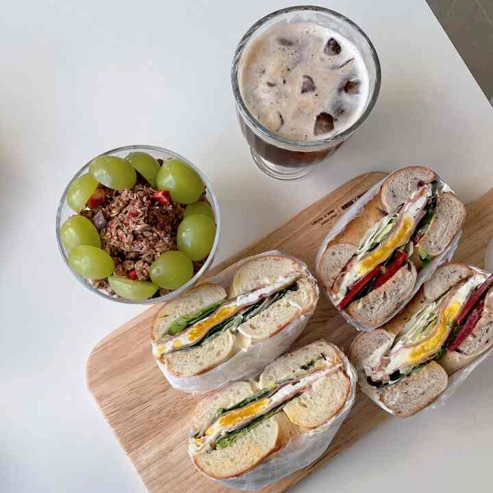 明るいホワイトと温かみのあるウッドトーンのワンルームキッチンテーブルに、ベーグルサンド、アイスコーヒー、ヨーグルトボウルが置かれたシンプルなおうちカフェの空間