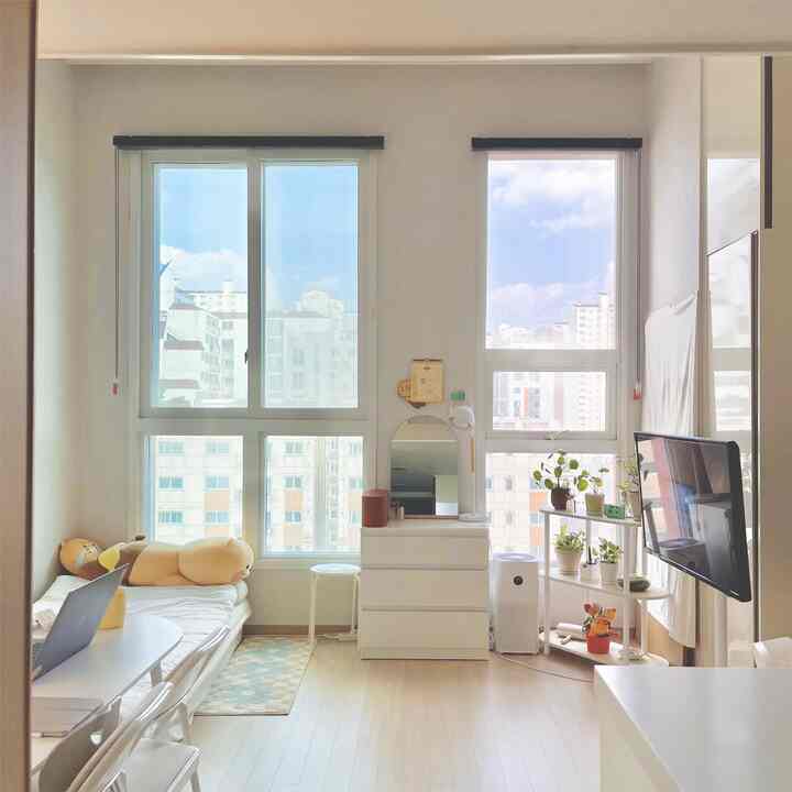 白とベージュのトーンの寝室、窓際に植物と棚、化粧台が配置された居心地の良い一人暮らしの空間