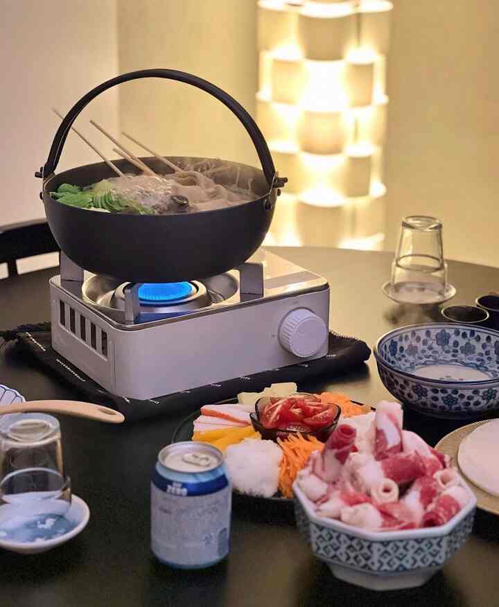 暖かな照明のダイニング空間、黒いテーブルにガスバーナーと鍋、多彩な料理が並ぶホームパーティー向けインテリア