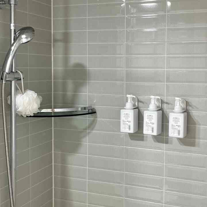 グレーのタイル壁、左側に白いシャワーヘッド、壁に取り付けられた3つの白いボトルがある清潔なバスルームの空間