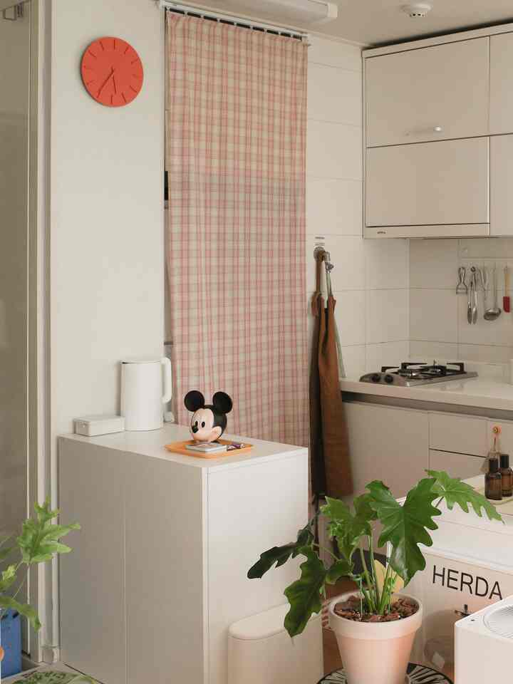 暖かいホワイトトーンとピンクのチェックカーテンがあるコンパクトなキッチン空間、緑の植物がアクセントとして配置されている