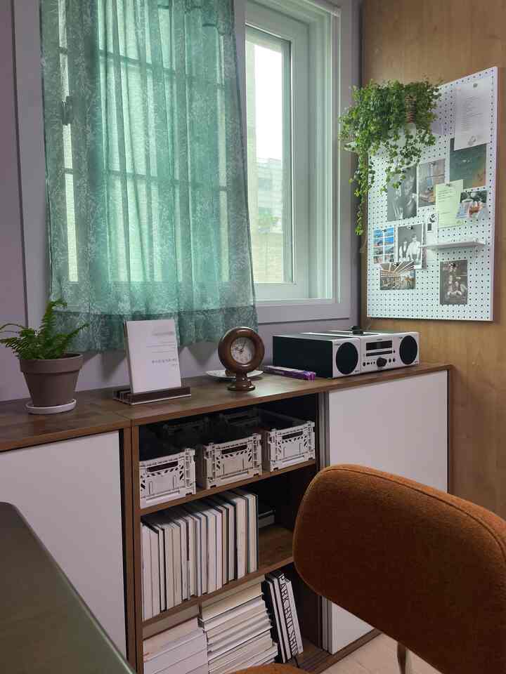 木目調と白色が調和した小さな部屋、収納キャビネットと窓、柄入りの緑のカーテンと写真が飾られた有孔ボードが配置された落ち着いた空間