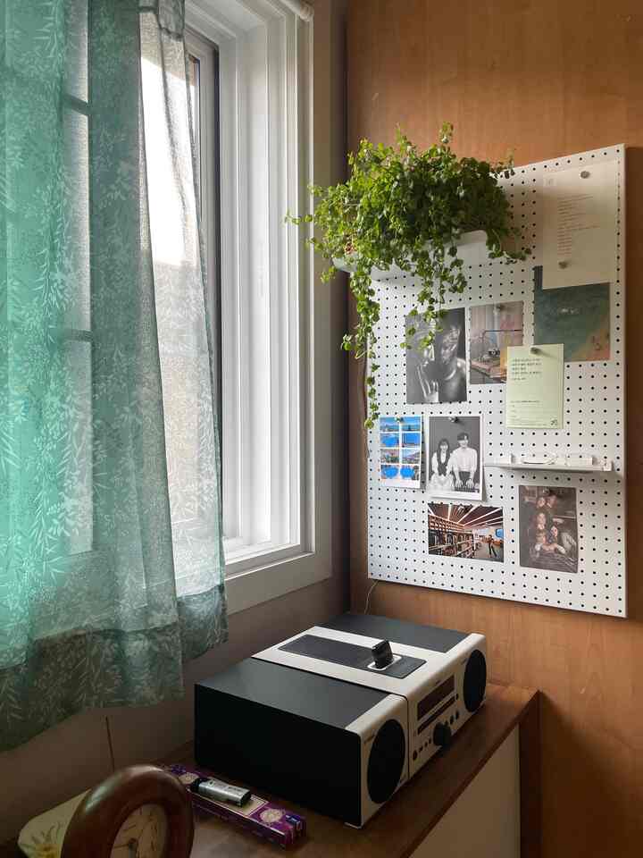 ナチュラルカラーのリビングの一角、ペグボードに写真と植物が飾られ、ブルートゥースオーディオとティールのカーテンが配置された印象的な空間