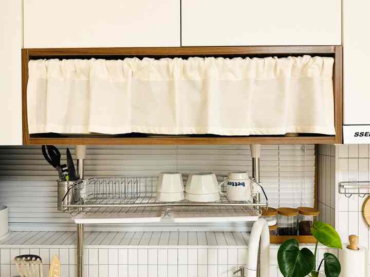 白と木目調が調和したキッチン空間、カーテンで覆われた上部収納と食器乾燥ラックが配置された雰囲気のあるインテリア
