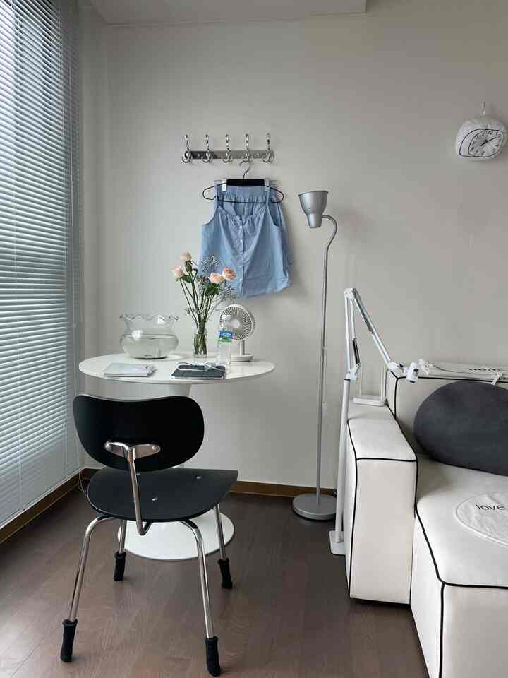 ホワイトとグレーのトーンの狭いリビング、シルバーのコートラックと丸いダイニングテーブル、ブラインドが配置された清潔感のあるインテリア