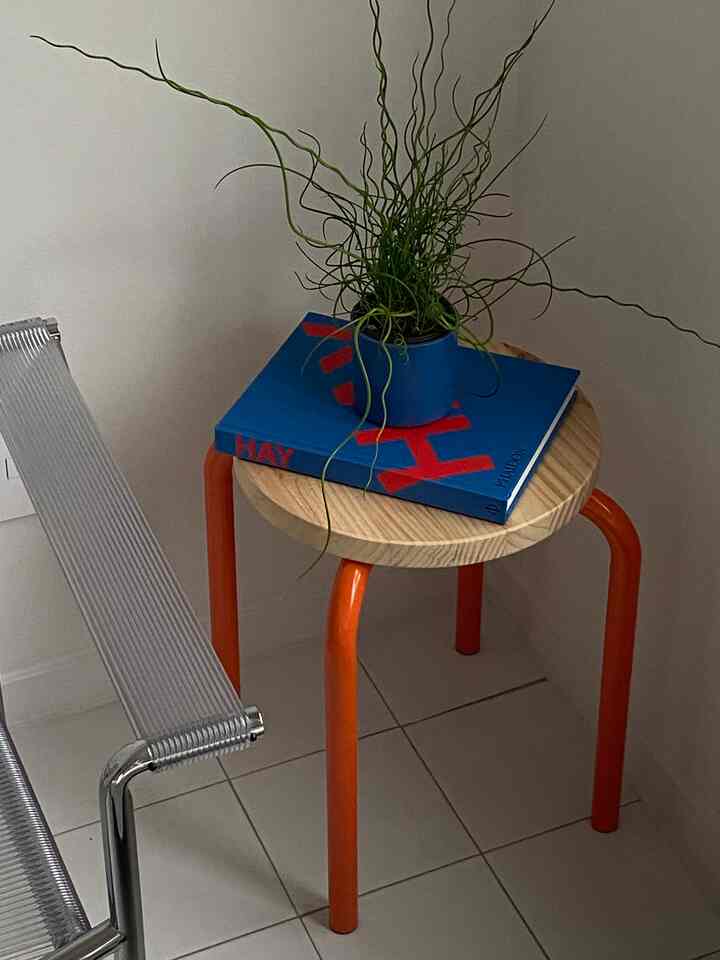 白いタイルの床と壁、オレンジ色の脚のスツールに木製の天板があり、青い本と緑の植物が置かれたモダンなキッチンの一角