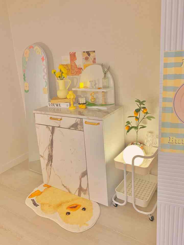 白と黄色のトーンのパウダールーム、マーブル模様のキャビネットとひよこのバスマット、デコステッカー付きの全身鏡が配置された居心地の良い小さな部屋