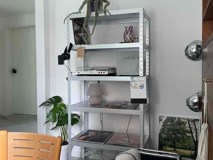 白い壁とメタルシェルフを配した狭い一人暮らしの空間、ターンテーブルやヴィンテージランプ、Bluetoothスピーカーがあるシンプルなインテリア