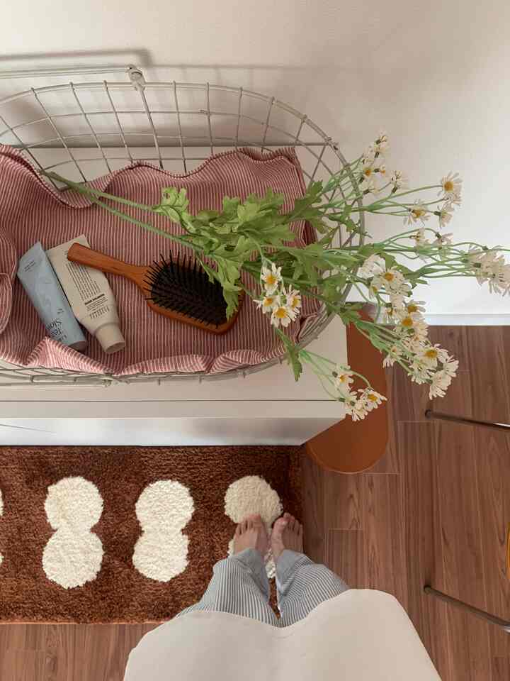 ブラウンの木製床と白い壁の空間、バスケットにハンドクリームとブラシ、花のオブジェ、ブラウン模様のラグが置かれた落ち着いた収納スペース