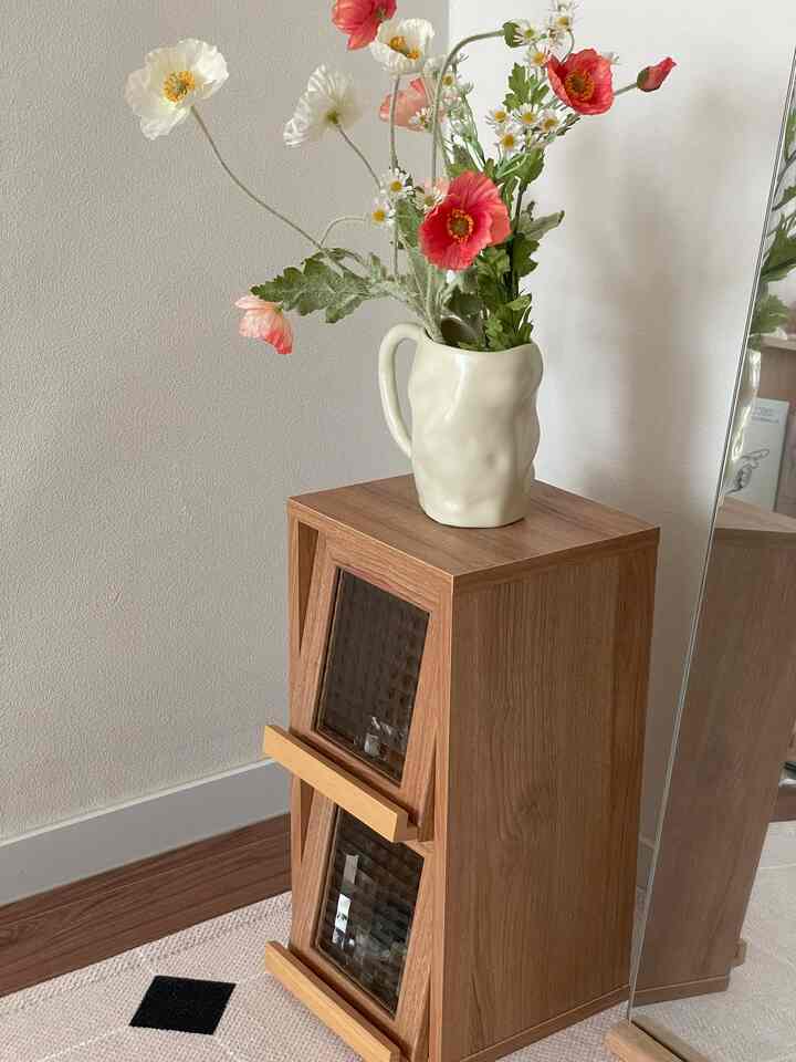 ナチュラルなブラウン色調の小型収納家具の上に、造花が入ったベージュの花瓶が置かれた清潔感のあるインテリア