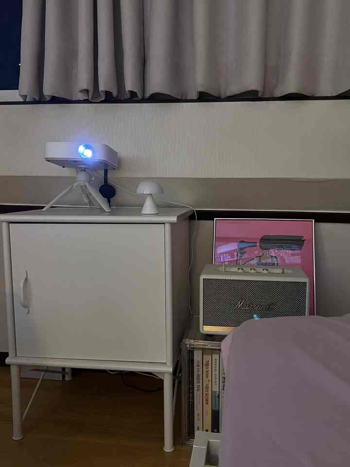 グレーの遮光カーテンとホワイトのナイトテーブル、プロジェクターとムードランプが配置された居心地の良い寝室