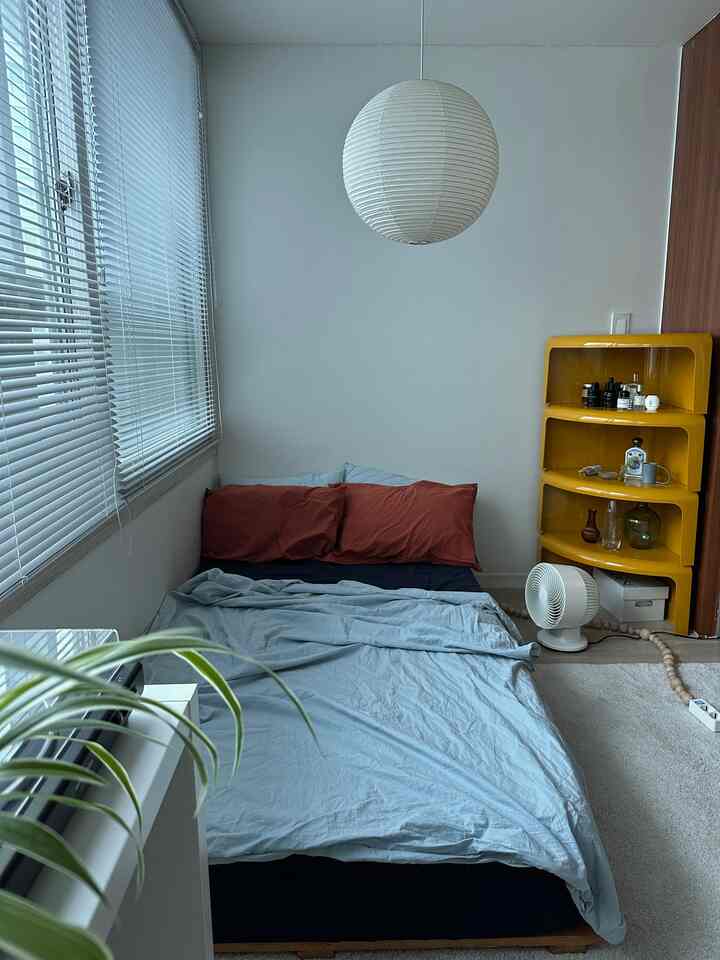 白色調の寝室、掛けられたアカリペンダントライトと黄色のコーナーシェルフが配置されたシンプルで居心地の良い空間