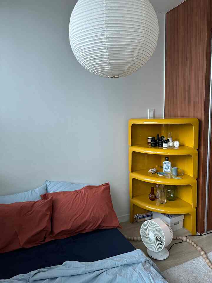 白と黄色を基調とした寝室、コーナーに黄色い棚とベッド、扇風機が配置された雰囲気のあるインテリア