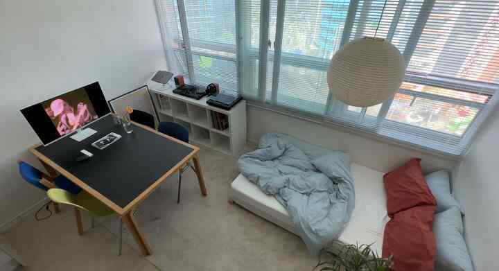 白い壁とナチュラルカラーの家具が配置された二人暮らし用のリビング兼寝室、ミッドセンチュリーモダンスタイルのダイニングテーブル、本棚、ベッドが見える空間