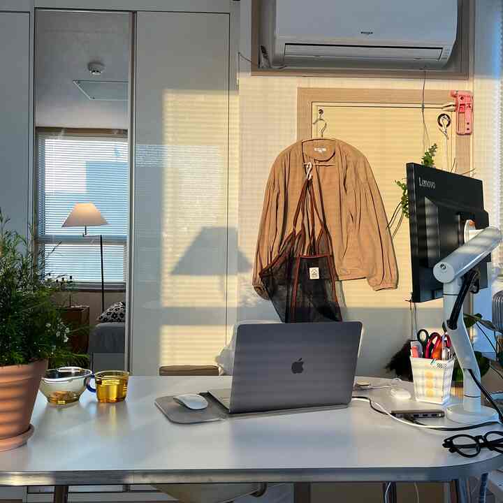 ホワイトとベージュのトーンのリビング兼ホームオフィス、白いデスクにノートパソコンとモニターアームが置かれ、暖かな自然光が差し込む空間