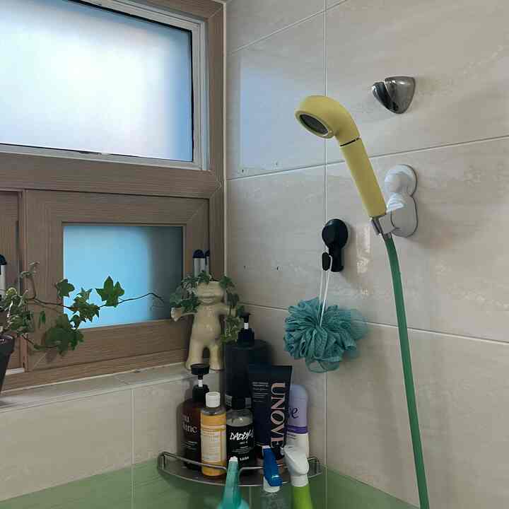 白と緑のタイル壁のバスルーム、黄色のシャワーヘッドと緑のホース、棚には様々な洗浄用品が置かれ、窓際には緑の植物が配置された雰囲気のあるインテリア