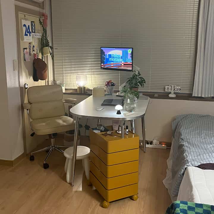 白とバター色を基調としたワンルームのリビング兼ワークスペース、中央に机と黄色いトロリーが配置された落ち着いたインテリア