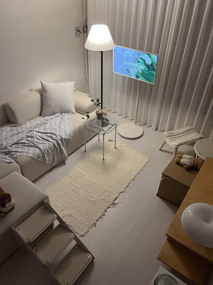 白とクリーム色調の暖かいリビング、ソファとフロアランプ、ペット用階段と猫が配置された雰囲気のある空間