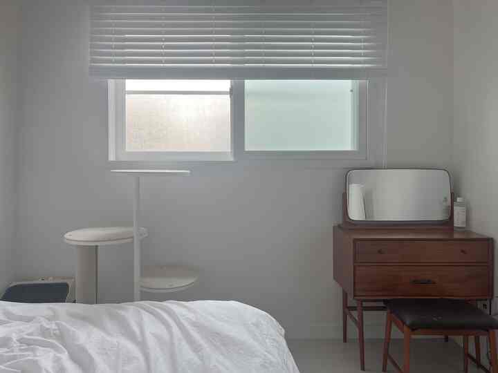 白とナチュラルな色調の寝室、窓のブラインド、キャットタワー、化粧台が配置された清潔感のある空間