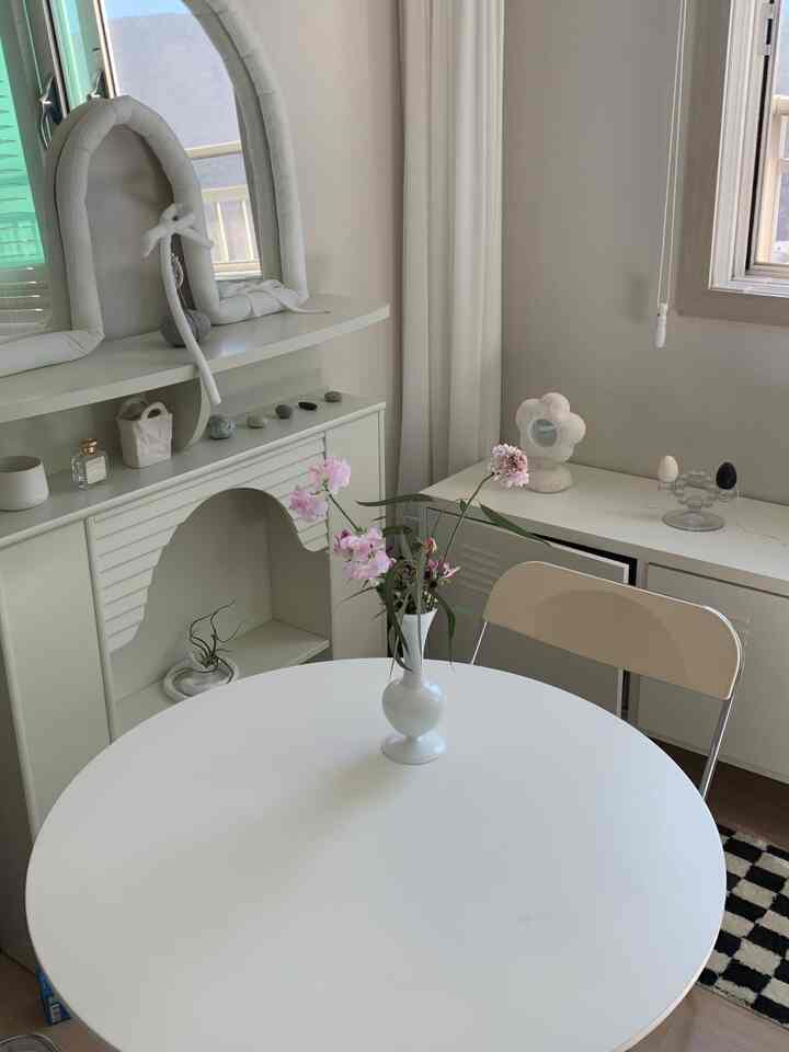 白色のトーンのダイニングルーム、丸いダイニングテーブルとキャビネット、花のあるシンプルな空間