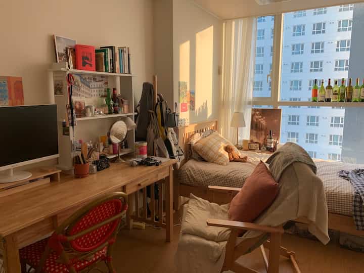 暖かいウッドトーンと白を基調としたワンルームの寝室、デスクとベッドが自然光の差し込む窓辺に配置された居心地の良い空間