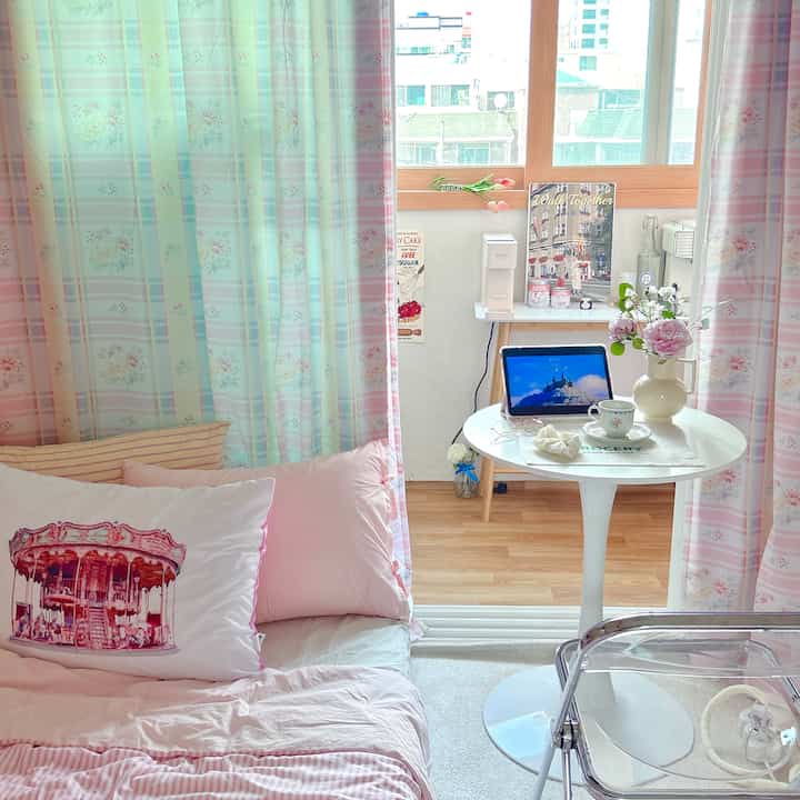 ホワイトとピンクのトーンが広がる小さな寝室、丸テーブルと可愛らしい花瓶が配置された雰囲気のあるインテリア