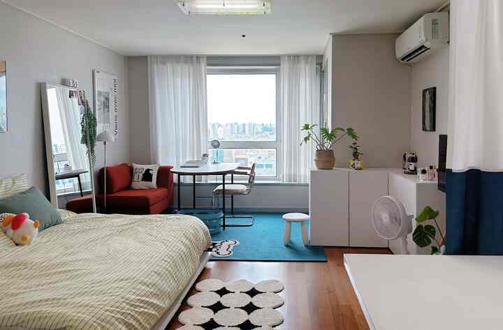 ナチュラルトーンと白いカーテンが調和するワンルーム、ベッドとソファ、デスクが配置された落ち着いた雰囲気の空間