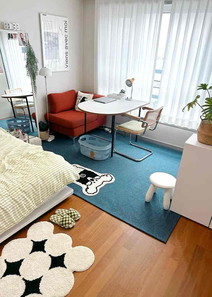 ネイビーのラグとオレンジの小さなソファが置かれた窓辺のシンプルな一人暮らし用ワークスペース兼寝室