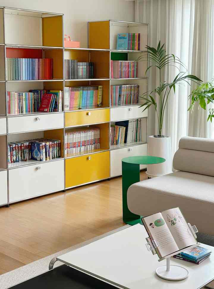 黄色と白のトーンのリビング、モジュール型の本棚と緑のサイドテーブル、植物が配置された清潔感のあるモダンな空間