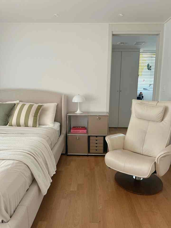 ベージュと白のトーンの寝室、ベージュのベッドとアームチェア、ナイトテーブルとテーブルランプが配置された自然で清潔感のある空間