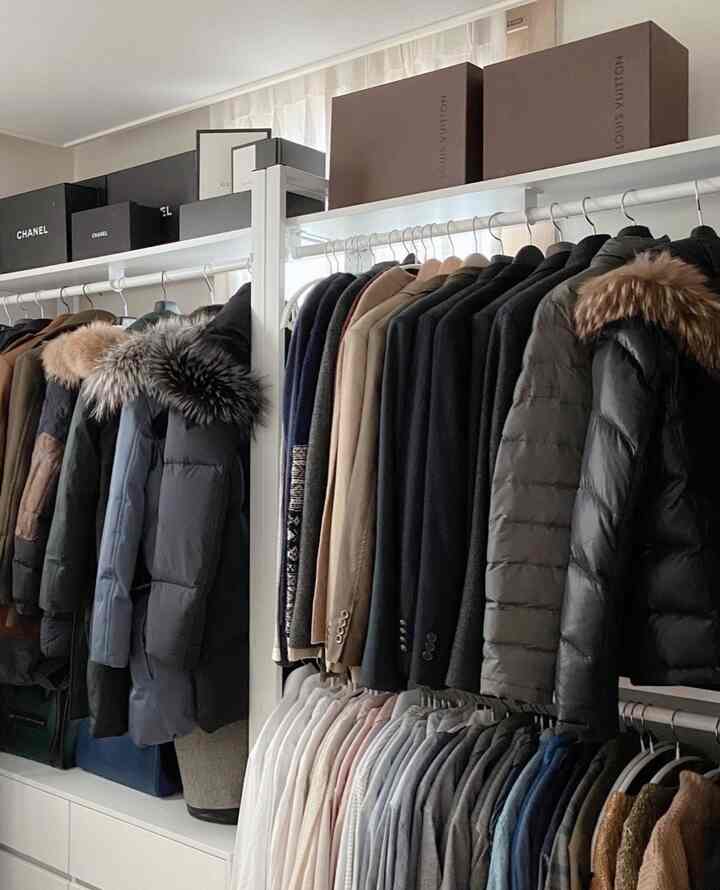 ニュートラルカラーのウォークインクローゼット、ベージュやグレー、ブラックの服が整然と並び、統一された白いハンガーと整理された収納棚が特徴の空間