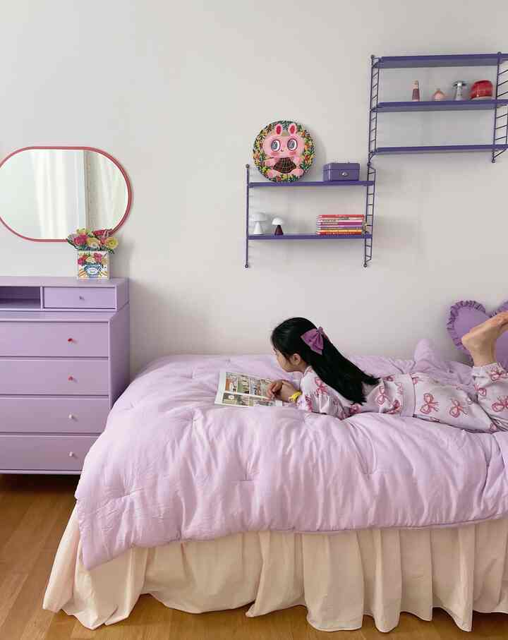 ラベンダー色調の子供部屋、ベッドとチェスト、壁の棚が配置された雰囲気のあるインテリアで子供が読書をしている様子