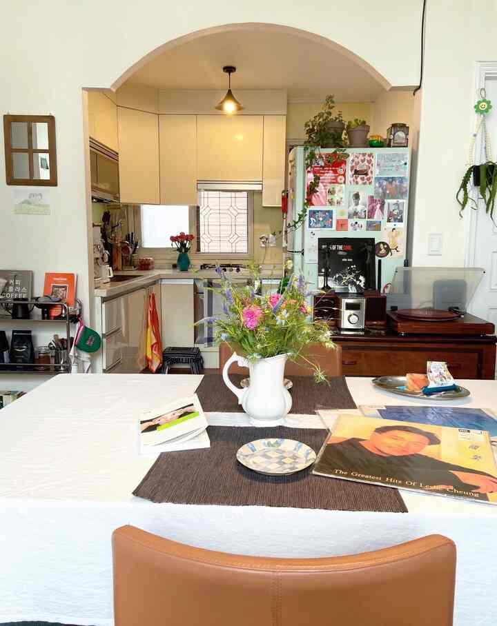 ナチュラルな色調のキッチンとリビング、中央に花瓶とテーブルランナーのあるダイニングテーブルが配置された落ち着いた雰囲気のインテリア