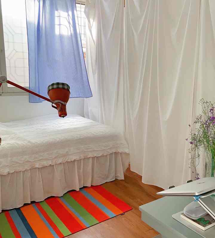 ホワイトとウッドトーンのコンパクトな寝室、フリル付き寝具とホワイトカーテン、多彩なストライプラグが特徴的なインテリア