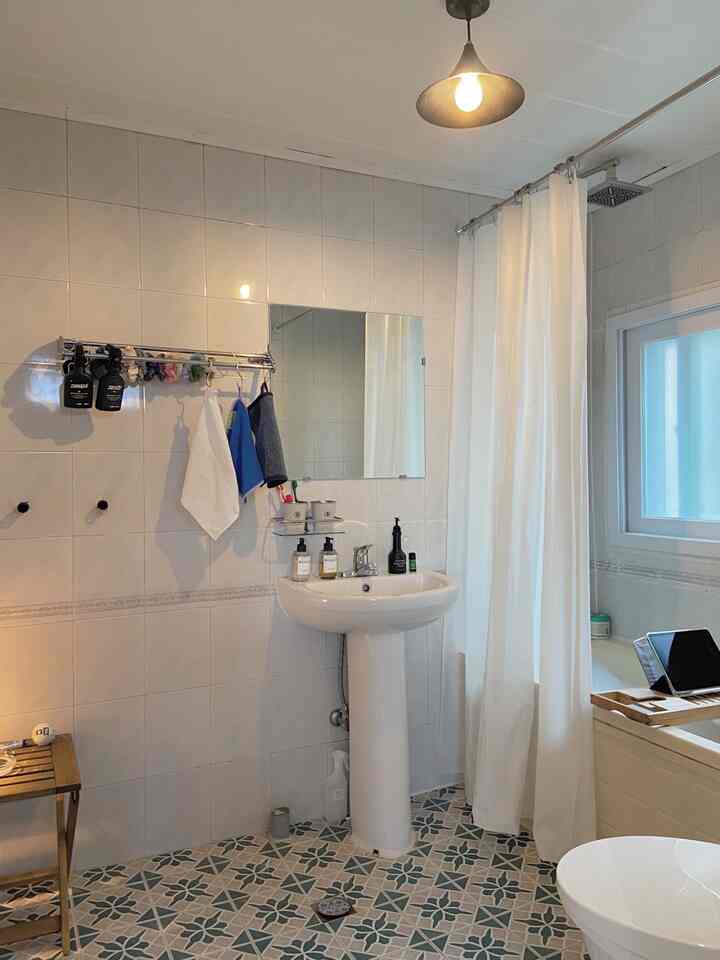 白とナチュラルな色調のバスルーム、洗面台とシャワーカーテン、木製スツールが配置された清潔で明るい空間