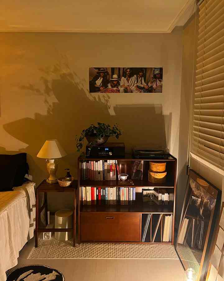 暖かい木目調とポスターが調和する居心地の良い寝室、書棚とスツール、テーブルランプが配置された自然なモダンなインテリア