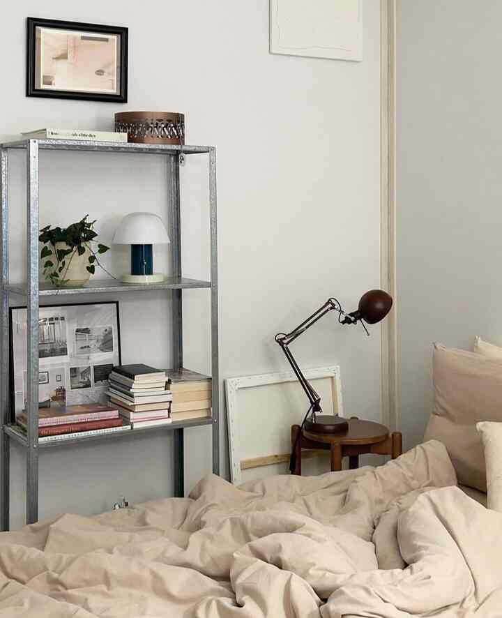 ナチュラルカラーの寝室、ベージュの寝具とメタル棚、木製スツール、テーブルランプが配置された雰囲気のあるインテリア