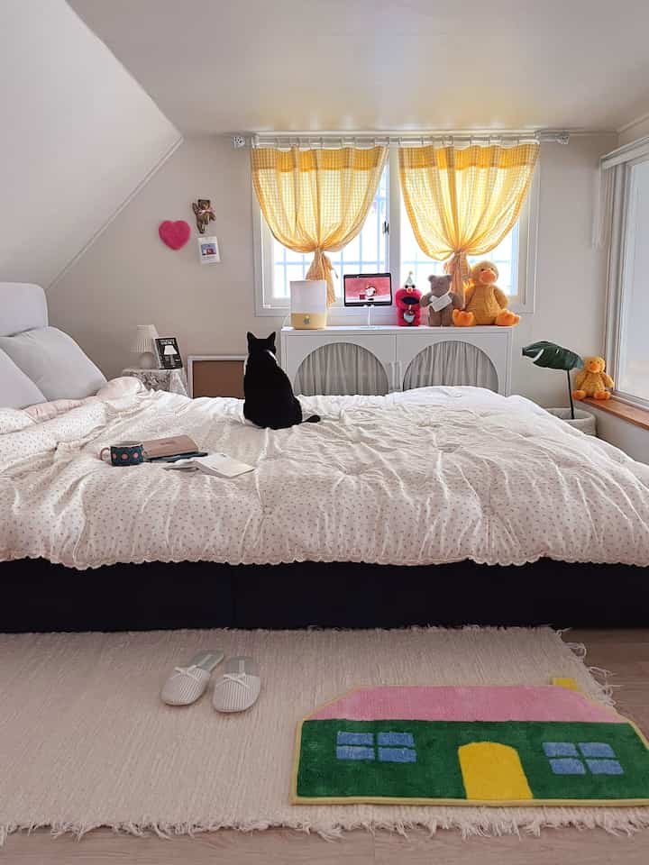 白と黄色のトーンの寝室、大きなベッドに黒い猫と黄色のチェックカーテンが配置された印象的な空間