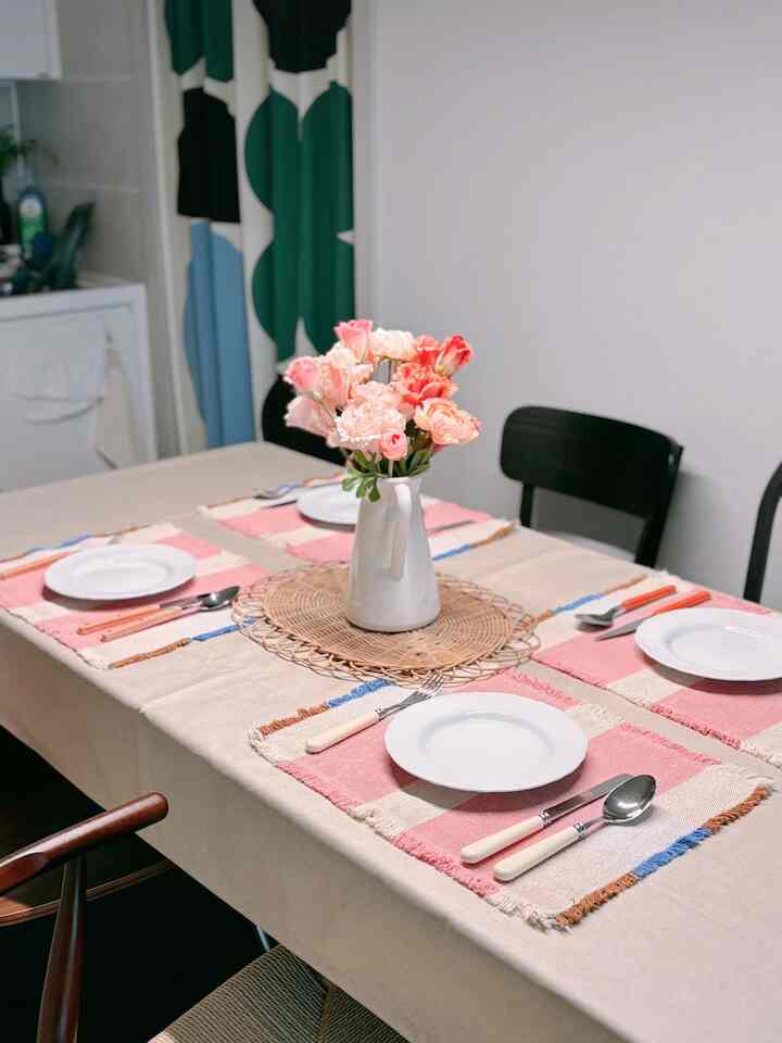 ブラウンの木製ダイニングテーブルにベージュのテーブルクロスが敷かれ、白い花瓶にピンクの花が飾られた温かみのあるダイニングルーム