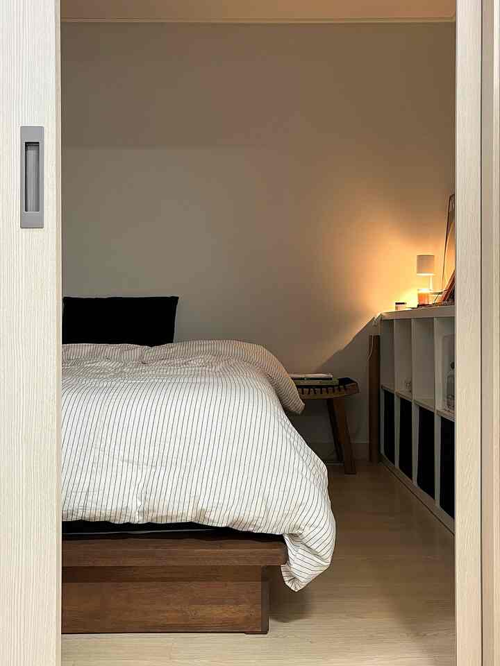 白い壁と木目調の床のコンパクトな寝室、ローベッドとストライプの掛け布団カバーが配置されたシンプルで落ち着きのあるインテリア