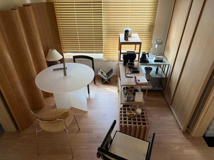 ベージュとウッドトーンのブラインドがあるリビング、透明な丸テーブルと多様な木製およびステンレスの棚が配置されたモダンで落ち着いた雰囲気のインテリア