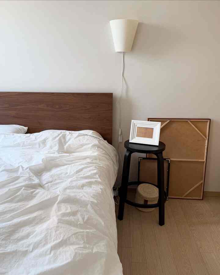 白い寝具と木目調のベッドヘッドがあるシンプルな寝室、黒いナイトテーブルとフロアランプが配置された落ち着いた空間