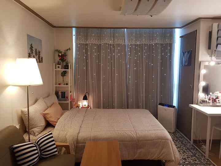 Beige-toned small bedroom featuring a central bed with blackout curtains, sofa and floor lamp on left, vanity and mirror on right, cozy atmosphere