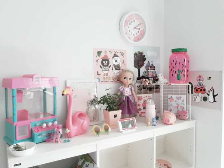 Pink and white toned kids' room featuring a shelf with cute toys and wall clock for a cozy atmosphere