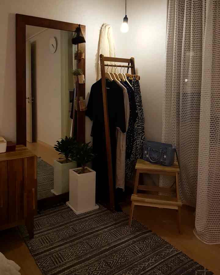 Light brown wood-tone bedroom space in front of a walk-in closet featuring a full-length mirror, wooden coat rack, stool, and plants creating a cozy atmosphere