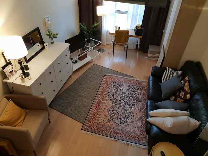 Cozy living room with brown wooden floor, black sofa, white dresser, rugs, and a desk with lighting by the window